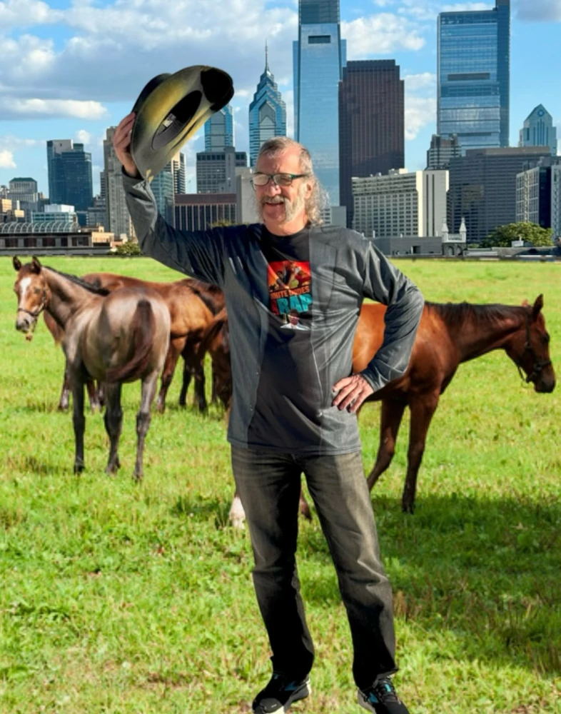 Hurley McNare holding a cowboy hat over his head while standing in front of horses in a field in front of the high rise buildings of Philadelphia.